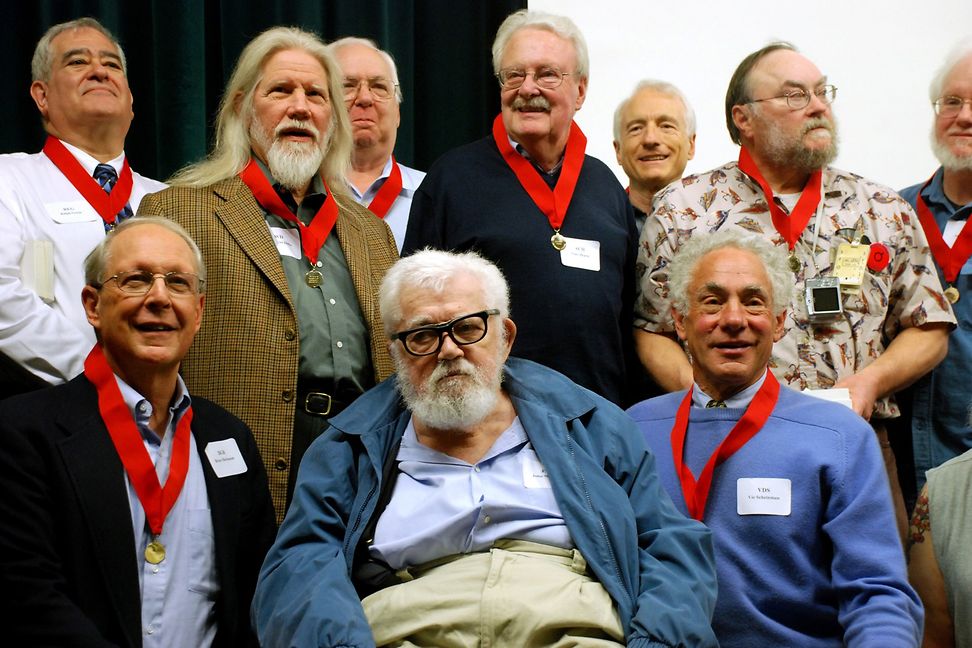 A group of elderly men with red lanyards around their necks pose for a group photo.