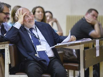 An older gentleman in a suit and tie sits in a lecture hall, resting his head on his hand and laughing