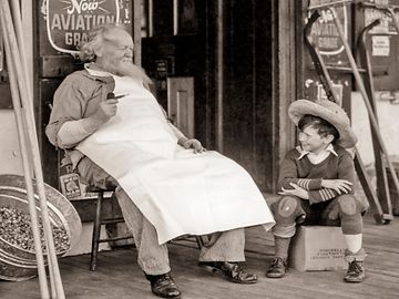 An elderly man in an apron sits on a chair in front of an old grocery store, next to a child perched on a crate.