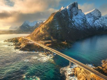 Aerial view of a bridge spanning a body of water, connecting a rugged, rocky landscape to a mountainous region.